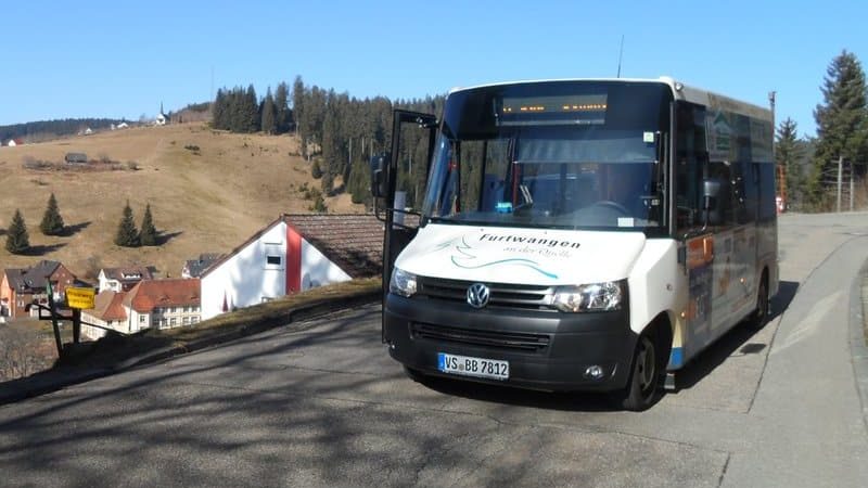 Ein weißer Kleinbus fährt auf einer offenen Landstraße. Man sieht der Bus mittels einer schrägen Vorderansicht. Es ist ein sonniger Tag mit blauem Himmel.
